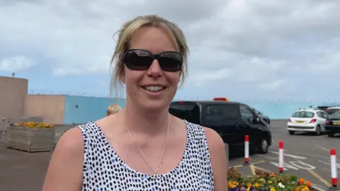 A woman stands in an outdoor parking lot, wearing a sleeveless white top with black spots and a silver necklace. Behind them are several parked cars, including a black van, and flower beds in wooden planters. Red-and-white striped bollards and a light blue wall topped with barbed wire are also visible under a cloudy sky.