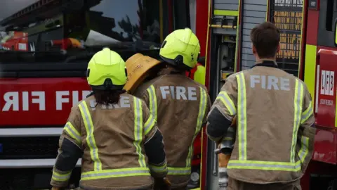 Three firefighters in uniform walking towards a fire engine with a hose