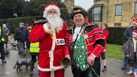 A man dressed as Father Christmas stands with the town crier, who wears a red and white ceremonial uniform with a green festive waistcoat underneath. Both are smiling at the camera. 