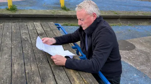 David Spereall/BBC A man in his sixties sits at a picnic table reading A4 sheets of paper. 