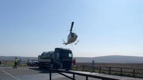 Helicopter hovering over a water bowser in a car park with moorland in the background