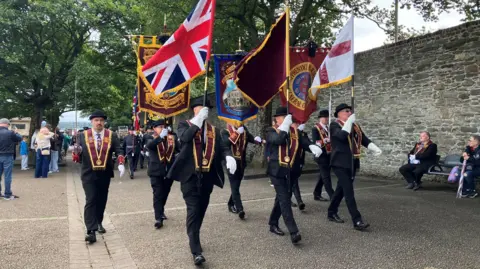 A group of men wearing black suits and black bowler hats marching in a parade. They are wearing maroon sashes an carrying various flags. There is a band behind them and old walls to their left.