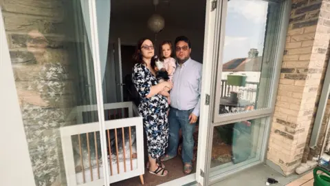 A couple holding a young child stands in their flat’s doorway beside a glass-panelled balcony.