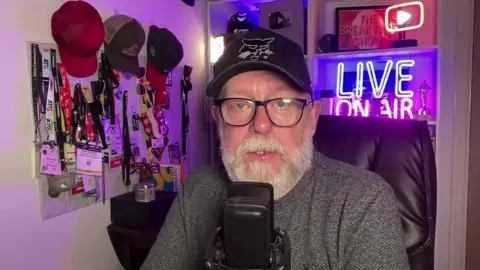 Demolition News editor Mark Anthony in his home studio, which speaks in front of a microphone. There are hats and lanyards on a wall next to him. Behind him there is illuminated YouTube -Long shield and live in the air sign. He looks into the camera.