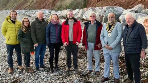 A picture of eight people pictured on the beach at Seaton. There is a rock pebble beach with a large rock and wall behind it.