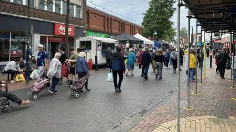 A market day in Bilston. There are lots of people walking the streets with shopping trolleys. We can see high street brands like Argos and Iceland.