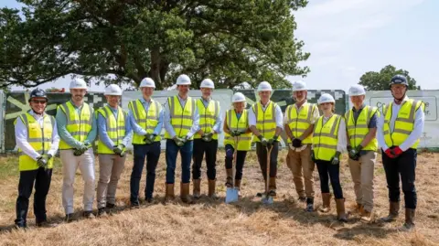 Twelve people, all in high vis jackets and hard hats, pose for a photo in a line. Two hold spades, while they all stand on brown grass, with a big oak tree in the background.