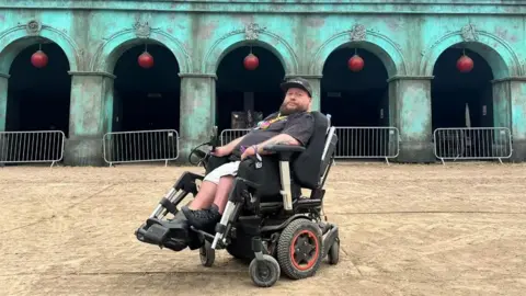 Karl Flood sat in his wheelchair outside of Rum Shack at Glastonbury Festival. It is a large green building with five arches and pillars at the front. The words Rum Shack are on the roof