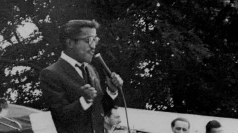 Sammy Davis Jr, dressed in a suit, sings on an outdoor stage with trees behind him