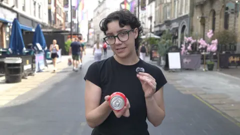 A woman with black rimmed glasses in a black t shirt is holding a red spherical plastic and metal drink called a BuzzBall in her right hand. In her left hand is a bluetooth microphone. In the background there are stone buildings that house a mixture of shops, bars and restaurants, there is also the rainbow themed pride flag and multiple people walking around the centre of Cardiff. There are also dark blue parasols and bright pink flowers on either side of the street decorating two of the aforementioned bars.