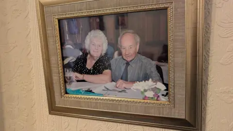 Alex Moss/BBC A framed picture hanging on the wall of a man and woman. The woman, on the left has short white hair and is wearing a black and white top. The man has short grey hair and is wearing a silver coloured shirt and dark grey tie. 