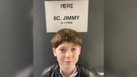 Albie Salter looking straight at the camera wearing a black leather jacket, standing in front of a black door with a white sheet of paper saying his character name "JIMMY".