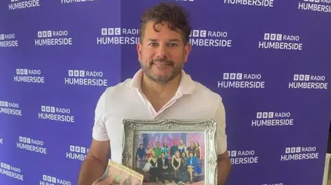 Zinc Grantham is standing in front of a purple-coloured BBC Radio Humberside wall. He has brown short wavy hair, a cropped beard and moustache. The hairdresser is wearing a pink T-shirt with a collar. He is holding a photograph taken with contestants of Blind Date and the host Cilla Black in 1997.