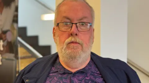 A man with blonde/grey hair and a beard looks into the camera. He has dark glasses. He is wearing a dark jacket and a purple and navy patterned shirt. In the background are stairs.