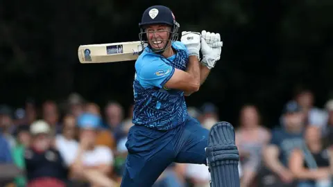 Wayne Madsen in blue Derbyshire Falcons kit holds his bat horizontally behind his head after playing a shot