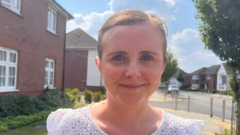 Young white female with brown hair in a tight pony tail. She wears a white short sleeved tshirt and is standing on a residential streets with trees nearby.