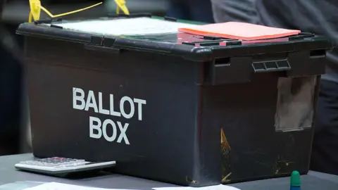 PA Media A black ballot box at a vote count. The words ballot box are written in white block capitals on the side of the box. A large calculator is next to the box, which is on a grey table. Pieces of orange paper are on top of the box.