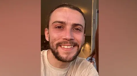 Callum Dickson A man with short brown hair and a brown beard and light brown jumper is smiling in this selfie.