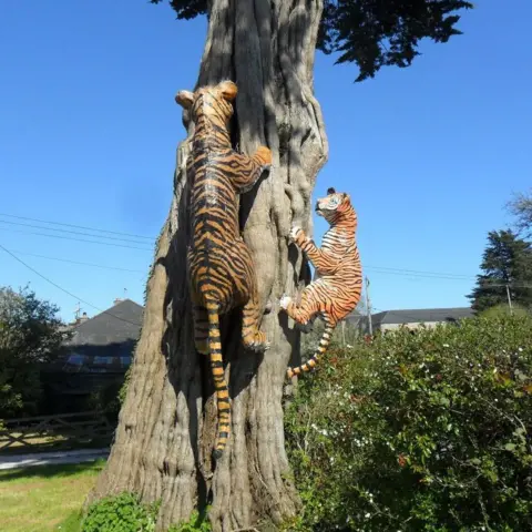 Malcolm Curley Two huge tiger sculptures are placed on a tree trunk as if they are trying to climb it. The location is on a farm with a big blue sky behind it.