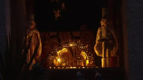 A window holds two large puppets stand either side of a small scene lit with yellow fairy lights which has a church and a stable and some figures.