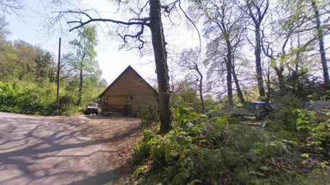 Google A single-storey garage-workshop with a high pitched roof on a narrow, winding country lane surrounded by tall trees.