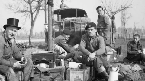 Getty Images A black and white picture of five soldiers looking towards the camera, surrounded by military kit. The man on the far left is wearing a top hat. They are resting by the side of the road. A sixth soldier stands on the roof of an amphibious truck in the background.