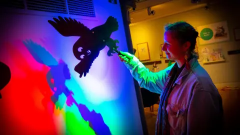The Late Shows A woman on the right holding a black cut out of an owl. She has her hair tied back and is wearing an oversized denim jacket and dangly earrings. Red, blue and green lights are being projected on the wall to the left, in a blue light. There is a wall behind the woman with various picture frames