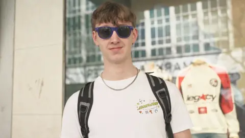 Oliver is pictured wearing a white graphic t-shirt, silver chain necklace and blue sunglasses. He has short brown hair which is longer on top and covers his forehead.