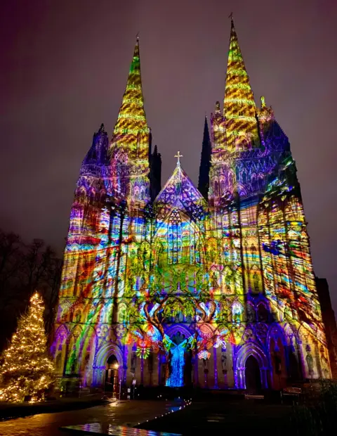 BBC Weather Watchers/Jeff Stevens The front of a cathedral at night with a bright, abstract, multi-coloured image projected on the front. A Christmas tree stands to the left.  