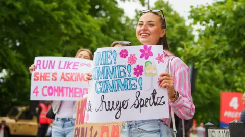 AJ Bell Great Bristol Run A young woman with dark hair, wearing a pink blouse and jeans, she is holding a motivational sign which reads Gimme, Gimme Gimme a huge spritz
