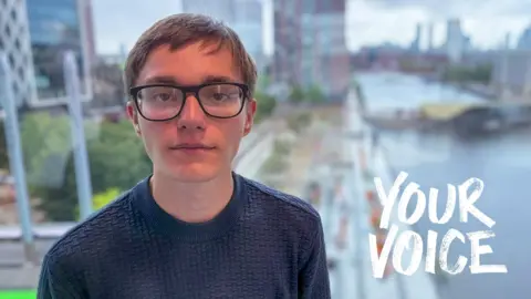 Dominic Hughes/BBC A young man wearing glasses looks into the camera with a blurred Salford docks in the background