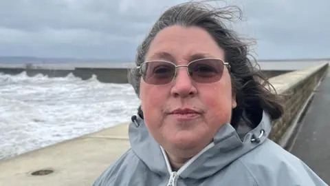 BBC Dawn Lord wears light brown sunglasses and her hair flutters in the wind. She is wearing a light gray coat, with the sea lapping against a seawall in the background.