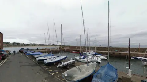 North Berwick Harbour showing a number of boats covered by the side of the water and several more in the water. Most of the boats are white in colour. The sky is overcast and the water is grey.