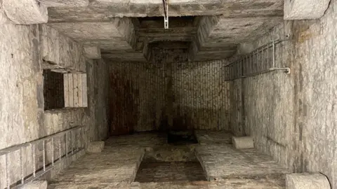 An underground stone passageway, deep and of some age. There are modern ladders on the left and right of the room. 