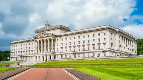 Getty Images The Northern Ireland assembly building. It is a large white square shape with many windows. There are five large stone pillars at the front door. 
