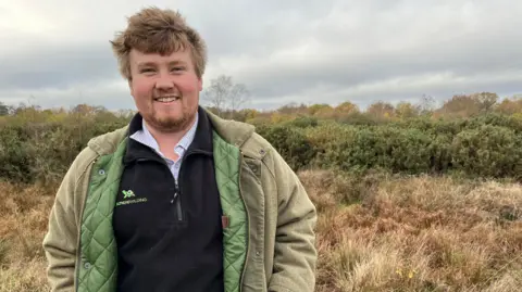 Clare Worden/BBC Harvey Tweats from Celtic Rewilding on the West Acre estate. Harvey has red/blonde hair and a beard. He is wearing a green coast with a black branded fleece under it. He is standing on grassland with gorse bushes behind him. 