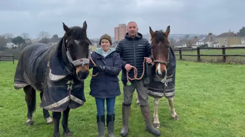 Sam Critchley (left) and Steve Hawkins stand in a field they both wear out door jackets, trousers and welly boots. They are leading a horse each. The horses are wearing outdoor rugs. A tower block and suburban housing are visible in the background.