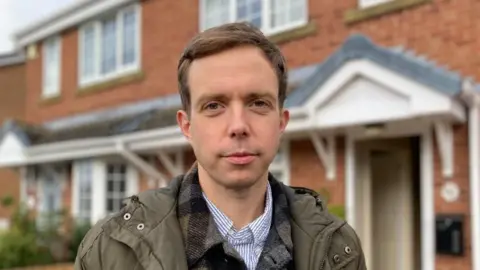 A man with short brown hair stands outside the redbrick houses wearing a green waterproof jacket, checked over shirt and a striped blue shirt.