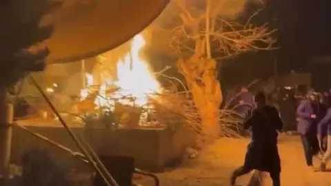 Phone footage Screenshot from phone footage, showing a satellite dish on fire with some people running past