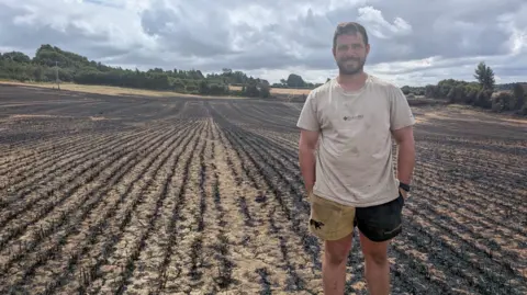 Newton Farm Farmer Josh Gay stands in a field damaged by fire. There are rows of blackened harvested crops behind him. He is wearing shorts and a t-shirt and has his hands in his pockets.
