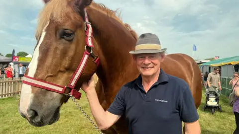 Actor Martin Clunes, wearing a grey trilby with a yellow band, and a blue polo shirt, is smiling and standing next to a large brown horse.