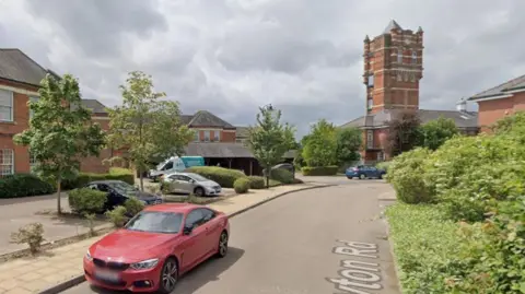 A Google image of Netherne-on-the-Hill village, with a red car on a road in the foreground and red brick buildings in the background