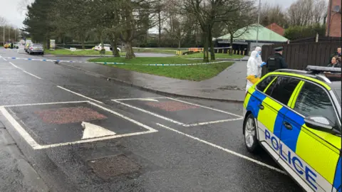 A police car in the right hand corner blocks a road alongside a green area. There is a police cordon, forensic officers in white overalls, and in the distance a white estate car. 