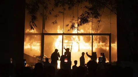 A fire can be seen in a building with a group of people silhouetted before the flames
