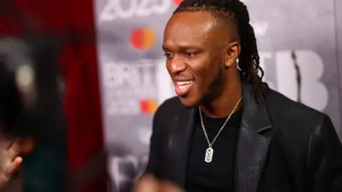 KSI is wearing a black blazer and t-shirt with a silver necklace.