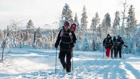 Beyond the Ultimate A woman in black arctic gear with black hiking poles is smiling. The hood is up and she has a pink neck scarf around her chin. There is a pink rucksack on her back with water bottles on the straps. She is hiking through dusty snow, and around her trees are covered in it, showing almost no bark. Behind her are three other people hiking 