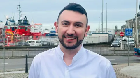 A man with dark hair and a dark beard smiles at the camera while standing in front of a harbour 