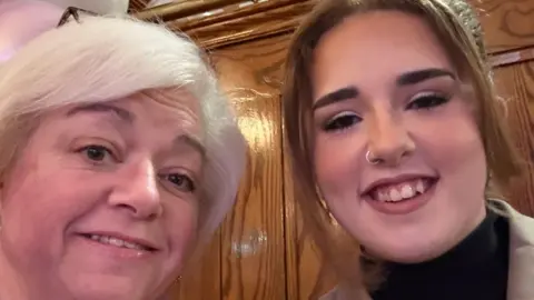 Andrea with short white hair. She is smiling. Ibbie has brown hair tied back and a nose ring. She has a beige jacket and black top. She is smiling.