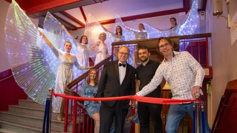 Ian Allington Photography / Westmorland and Furness Council The red ribbon reopening the venue is cut by councillor Steve Bavin, watched by new leaseholders Bryn Tyson (centre) and Mark Naunton. They are standing on a stairwell in the building and smiling at the camera. Dancers in glowing costumes are lined up on the top steps.