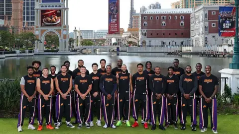 Getty Images A team photo of the Phoenix Suns basketball team by a body of water with a skyline of large buildings showing advertisements for their games with the Brooklyn Nets behind them. The team are all wearing black tops with the words Suns on the front in orange text and black trousers with yellow, orange and purple lining on them and the Phoenix Suns logo.